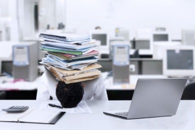 Man with his head on the desk under a large stack of papers. 