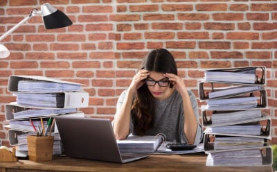 Woman at desk with head in hands surrounded by large stacks of documents. 