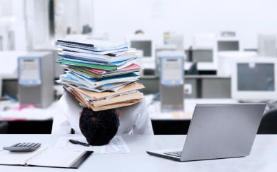 A man with his head on the table under a large stack of papers. 
